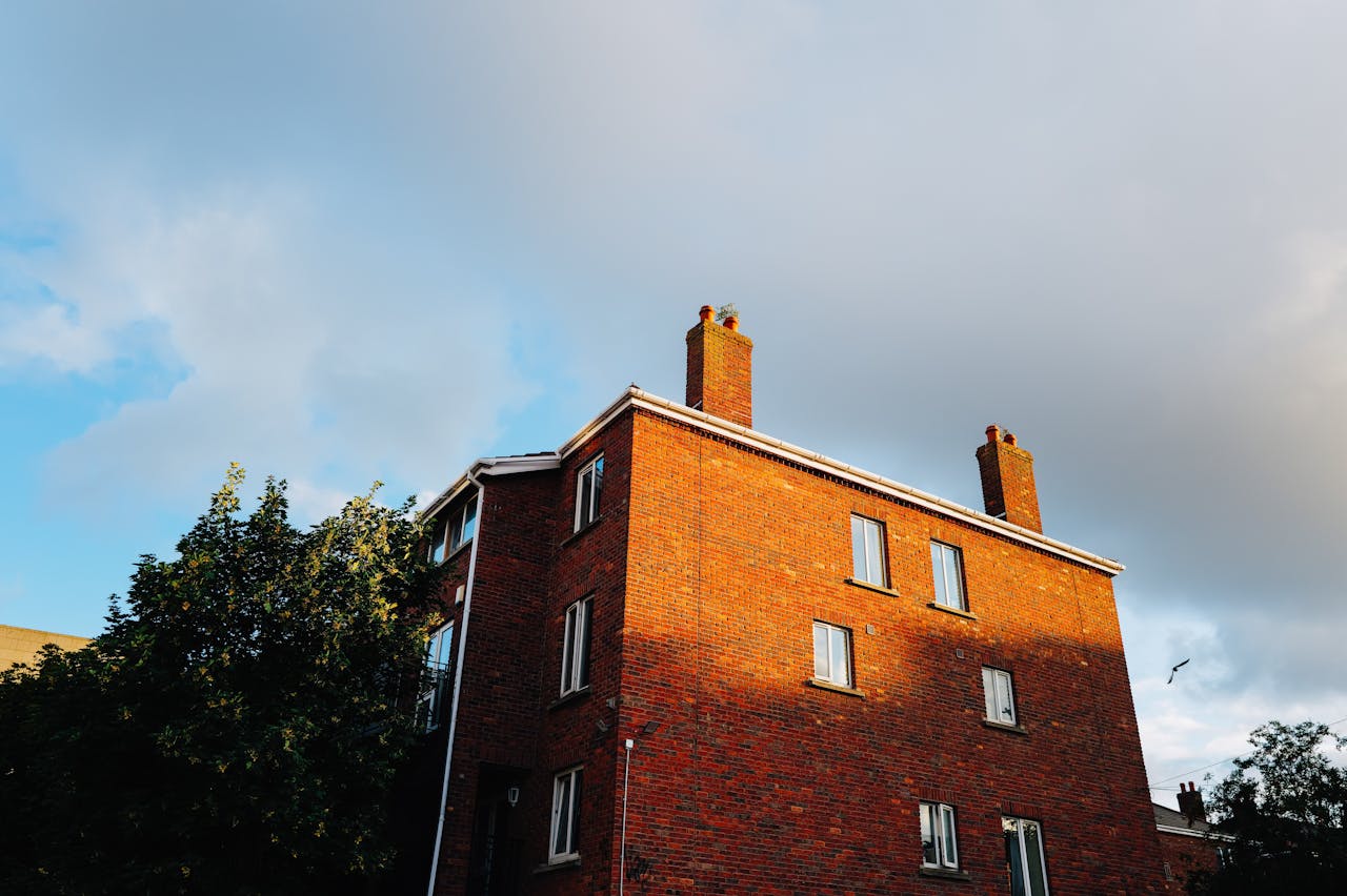 A red brick building with chimneys highlighted by sunlight against a partly cloudy sky.